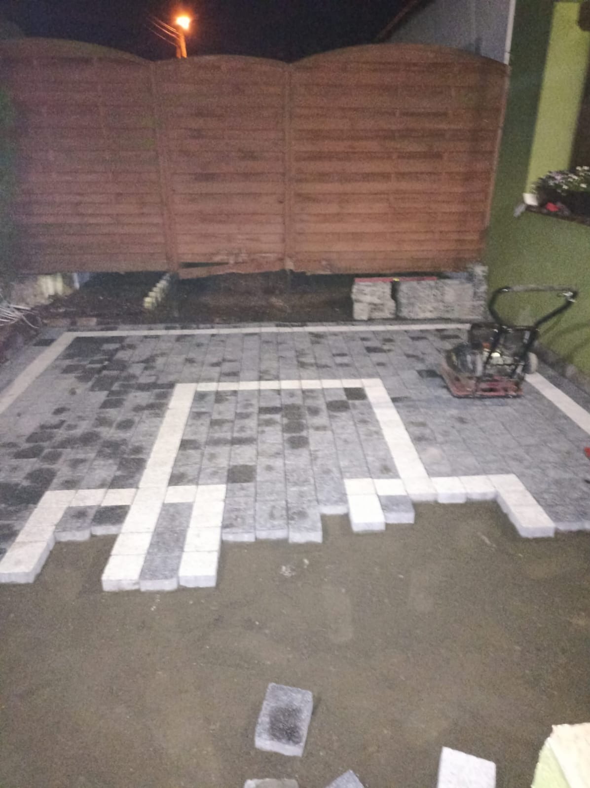 Układanie kostki brukowej w ogrodzie, widoczny fragment ułożonej nawierzchni z szarych i białych kostek, zagęszczarka w tle, etap prac.