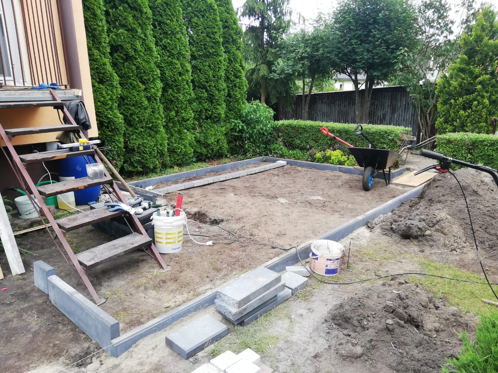 Przygotowanie podłoża pod taras: obrzeża z kostki, ziemia wyrównana, narzędzia pracy widoczne. Etap budowy tarasu w ogrodzie, widok z góry.