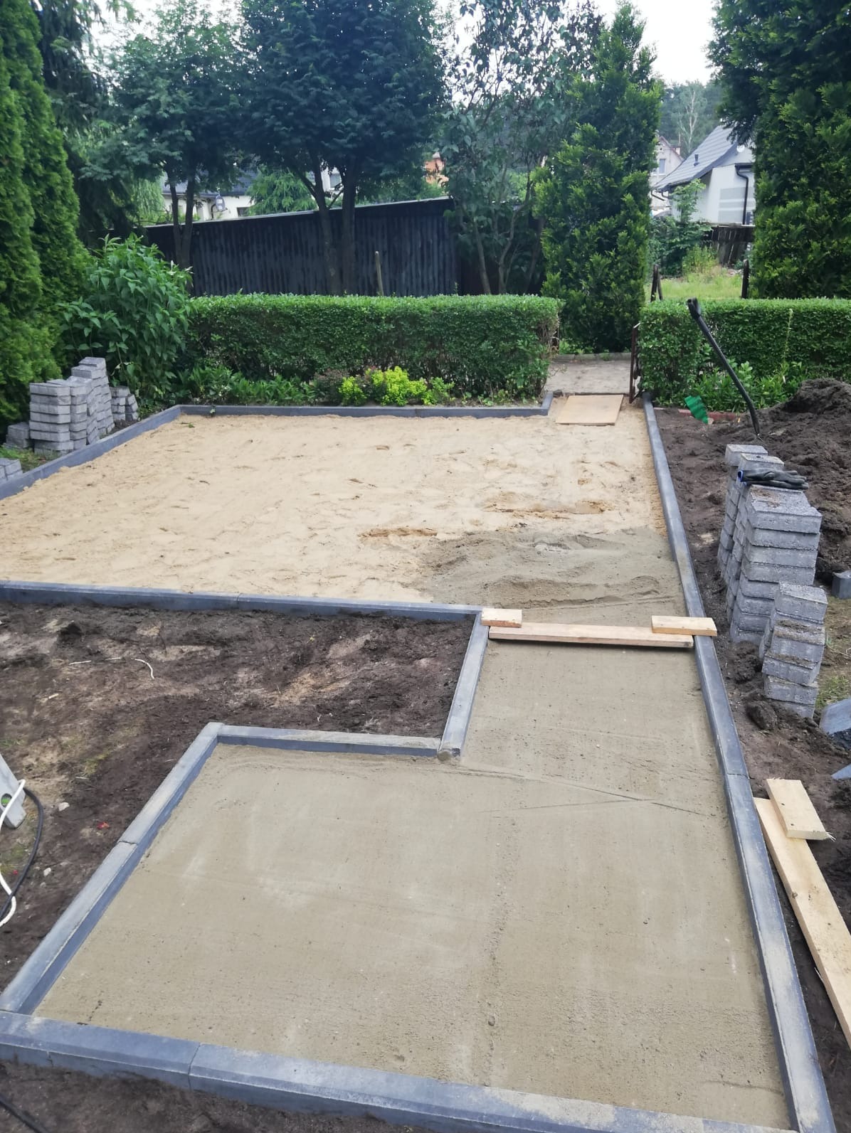 Przygotowanie podłoża pod kostkę brukową: obrzeża ułożone, piasek i podsypka wyrównane. Widoczne zapasy kostki i narzędzia. Prace ziemne w ogrodzie.