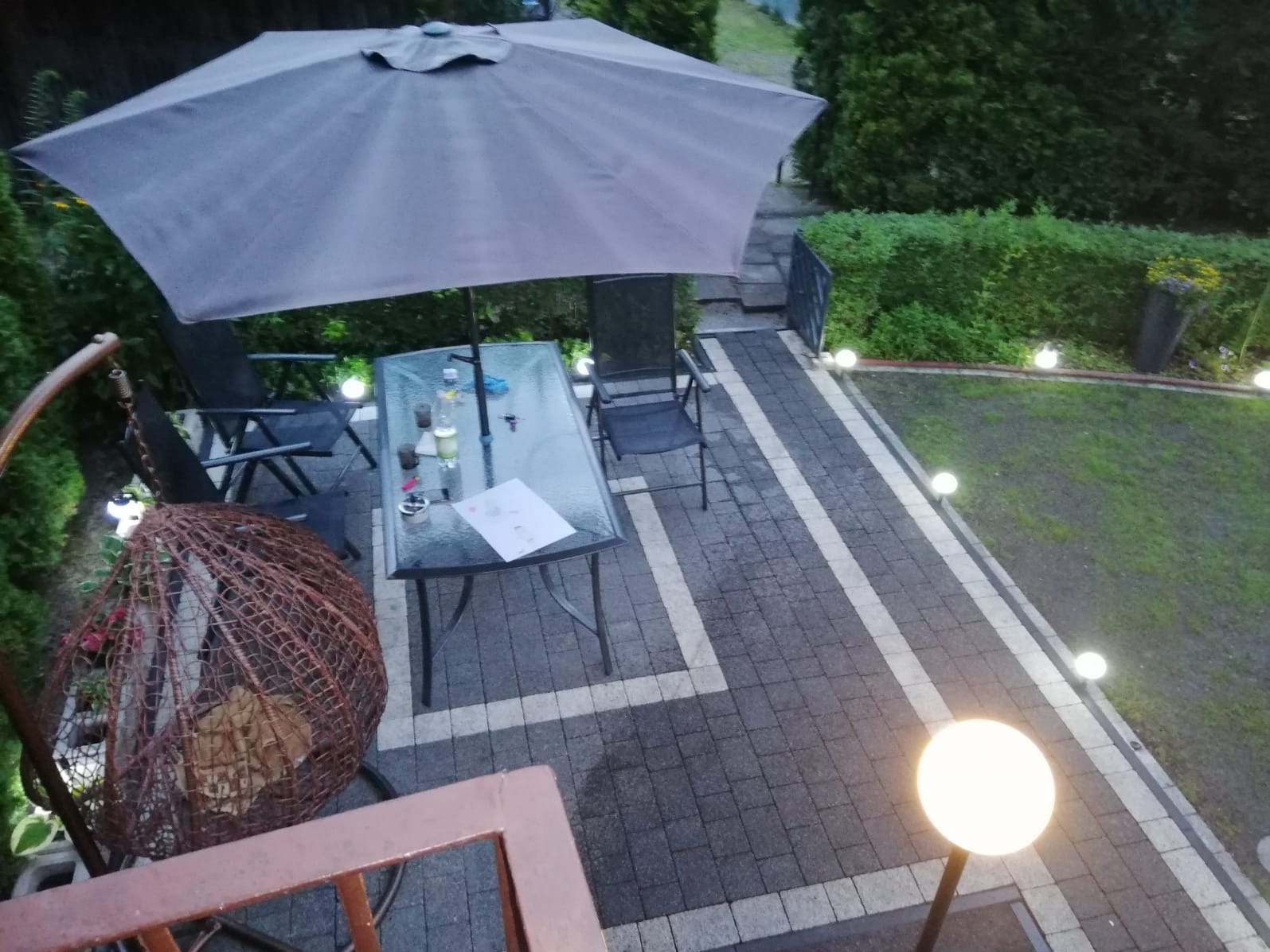 Oświetlony taras z kostki brukowej w szarościach, z meblami ogrodowymi pod parasolem. Widoczne lampy kuliste wzdłuż ścieżki i zielony trawnik. Na stole leżą dokumenty.