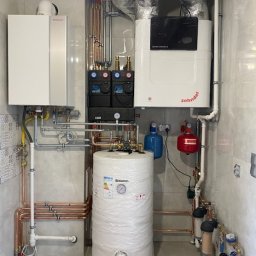TECHMAR - Złożona instalacja grzewcza z dwoma kotłami i zasobnikiem ciepłej wody w pomieszczeniu wykończonym płytkami. Widoczne liczne rury miedziane i elementy sterujące.