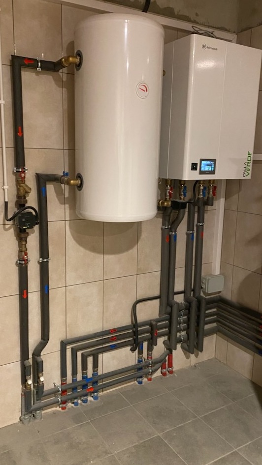 Biały bojler i pompa ciepła Dalla Verde z szarymi rurami w kotłowni wyłożonej płytkami. Instalacja z zaworami i oznaczeniami kolorystycznymi.