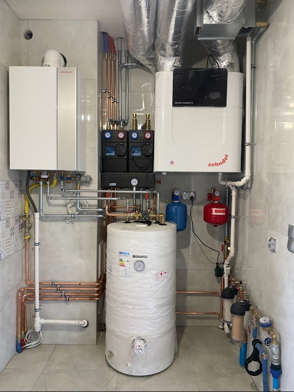 Złożona instalacja grzewcza z dwoma kotłami i zasobnikiem ciepłej wody w pomieszczeniu wykończonym płytkami. Widoczne liczne rury miedziane i elementy sterujące.