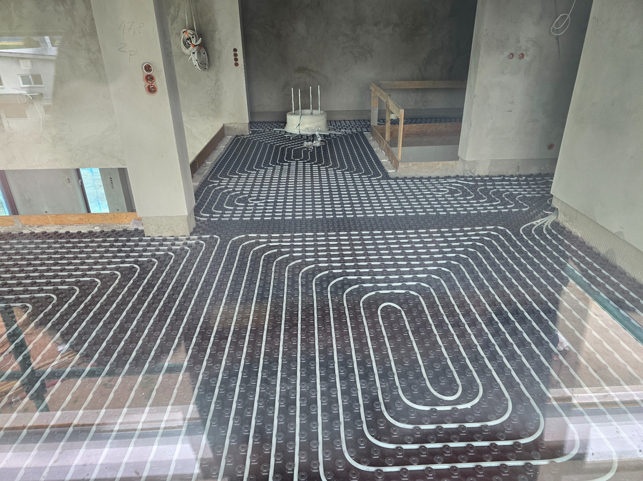 Ułożone rury ogrzewania podłogowego na czarnej macie w surowym wnętrzu. Widoczne białe rury w regularnych pętlach, przygotowane do zalania posadzką.