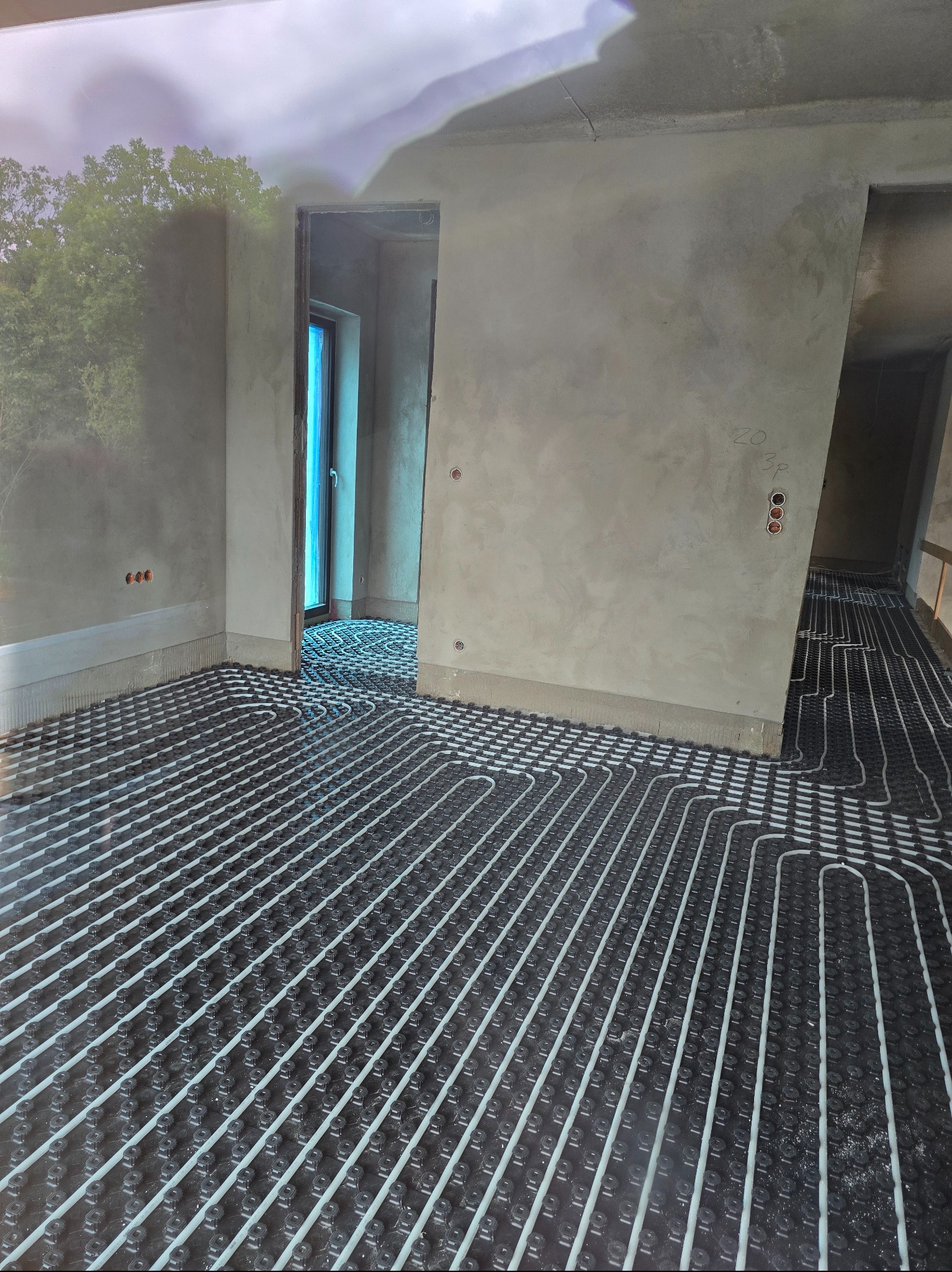 Ułożona instalacja ogrzewania podłogowego w surowym wnętrzu. Białe rurki na czarnej macie, widoczne otwory w ścianie i przejście do kolejnego pomieszczenia.