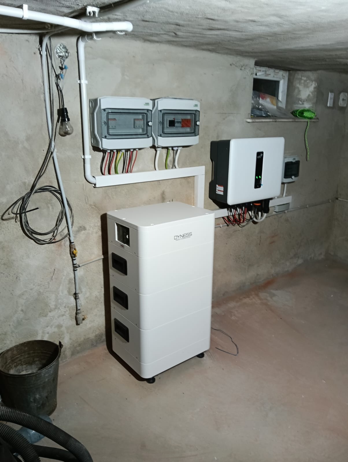 Zestaw fotowoltaiczny w piwnicy: biały akumulator Dyness, inwerter, rozdzielnice elektryczne z okablowaniem. Instalacja w surowym pomieszczeniu.