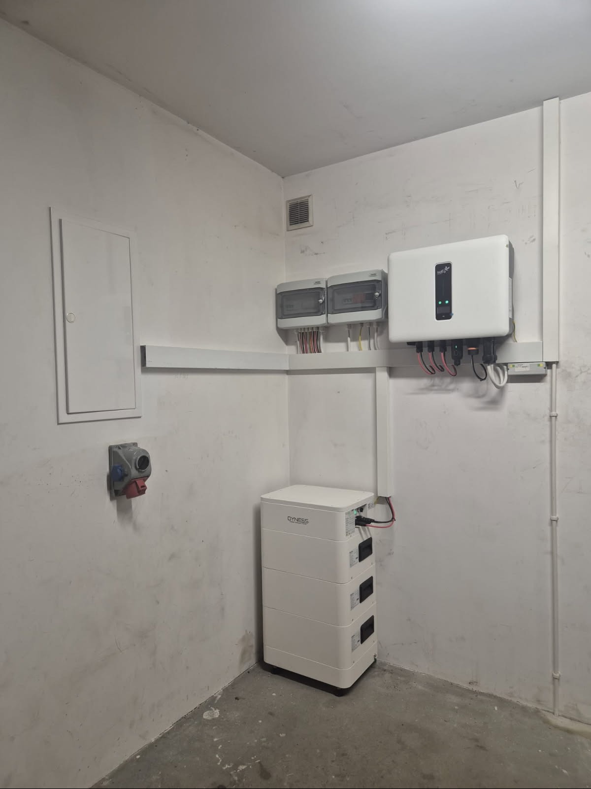 Biały akumulator energii Dyness, inwerter Sofar Solar i skrzynki elektryczne zamontowane na ścianie w pomieszczeniu. Instalacja fotowoltaiczna wewnątrz budynku.