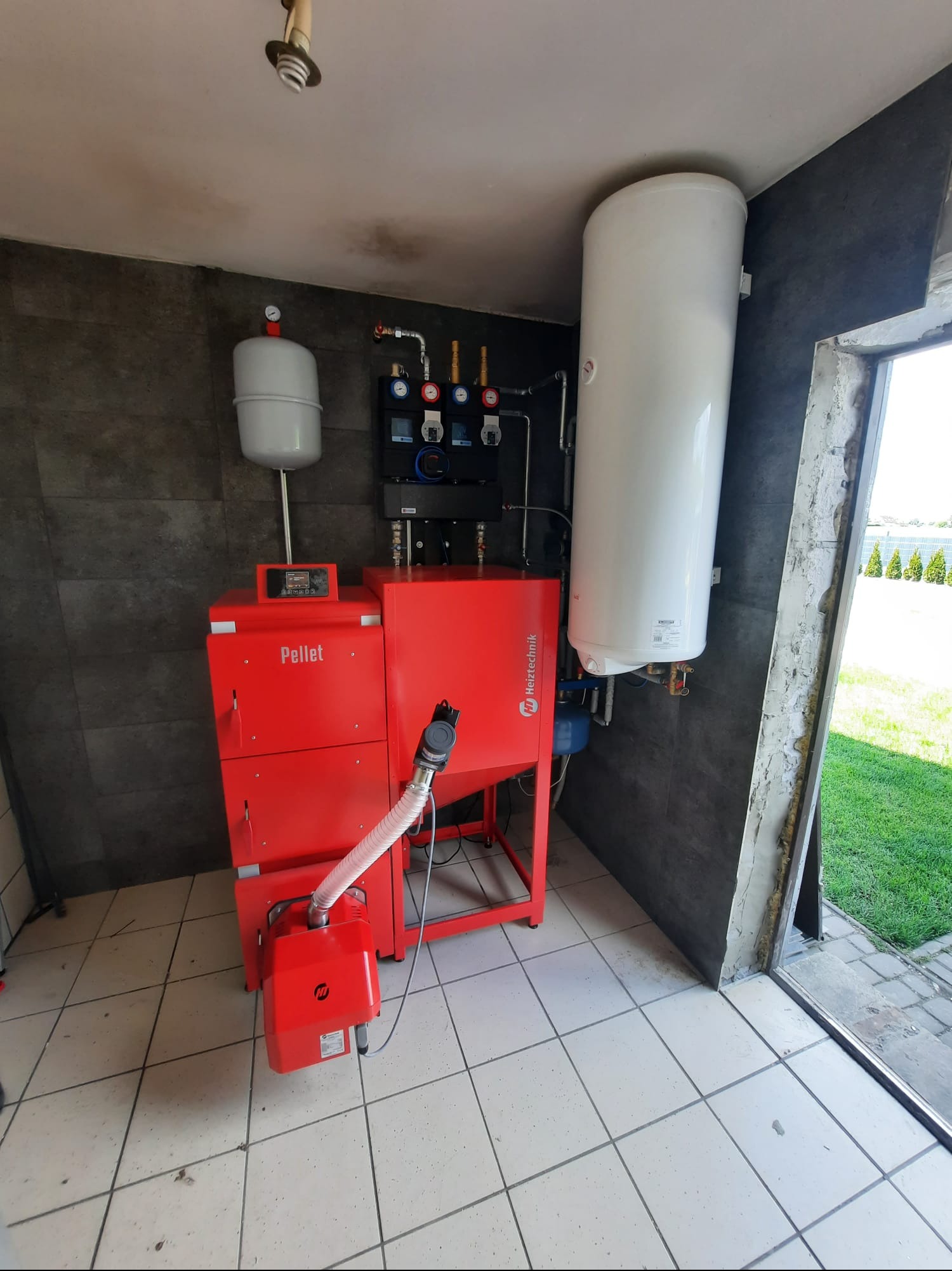 Czerwony piec na pellet Heiztechnik z zasobnikiem wody w kotłowni wykończonej szarą płytką. Widoczne elementy instalacji i fragment zieleni za oknem.