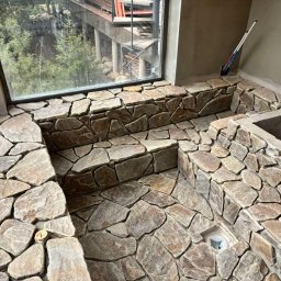 Adaro - Wykończona kamieniem łazienka z dużym oknem i widokiem na zieleń. Kamienna mozaika na ścianach i podłodze, odpływ liniowy. W tle widoczne narzędzia i elementy instalacji.