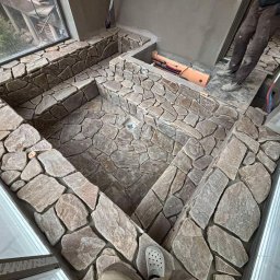 Adaro - Kamienna mozaika w łazience, ułożona z nieregularnych, szarych kamieni. Widoczne okno i fragment instalacji. Narożne ujęcie podkreśla unikalny wzór i fakturę.