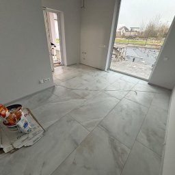 Adaro - Jasne, marmurowe płytki podłogowe w nowym wnętrzu. Widoczne wiaderka z zaprawą i narzędzia. Duże okno z widokiem na zewnątrz. Minimalistyczny styl wykończenia.