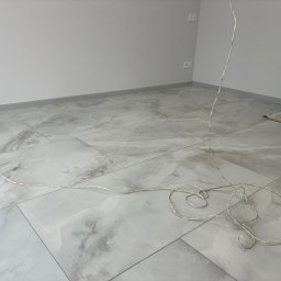 Adaro - Jasne, marmurowe płytki podłogowe w nowym pomieszczeniu z szarą listwą przypodłogową i zwisającym przewodem. Widoczne gniazdka elektryczne i akcesoria malarskie.