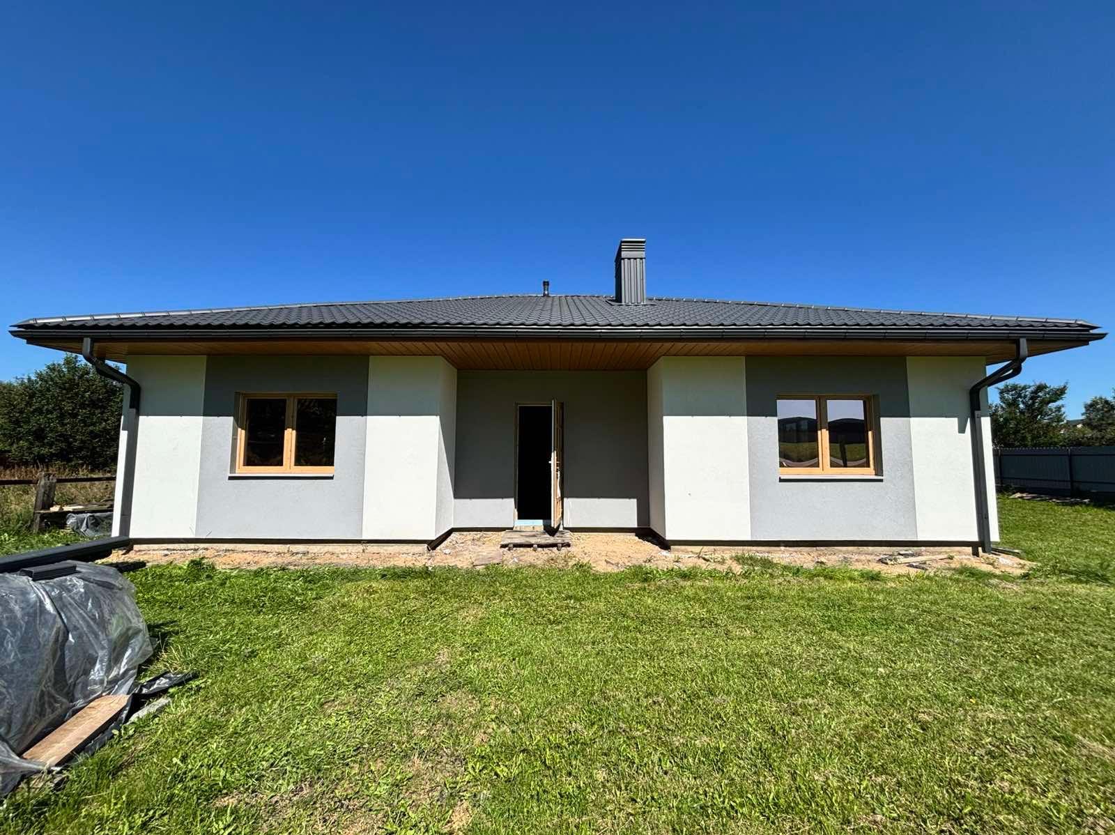 Nowoczesny dom parterowy z szarą dachówką i jasną elewacją w odcieniach szarości i bieli, z drewnianymi ramami okiennymi, na tle zielonego trawnika i błękitnego nieba.