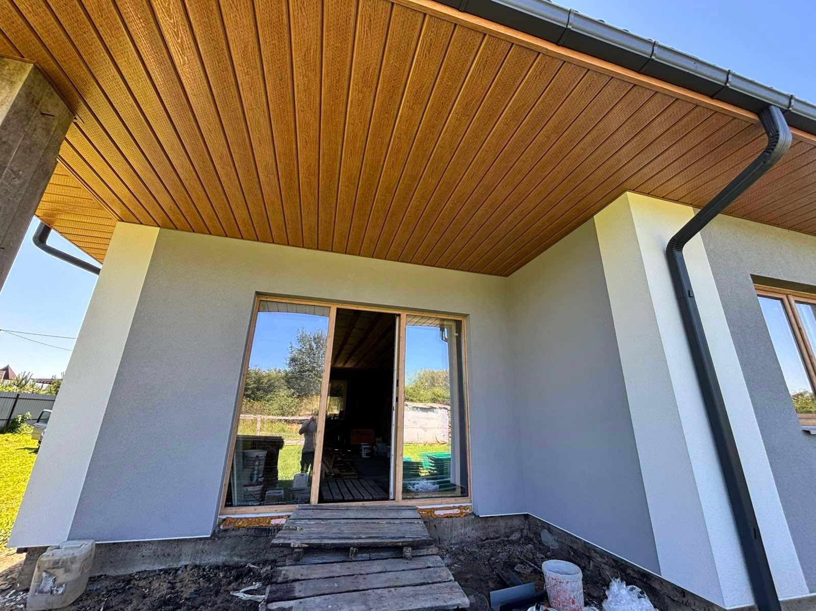 Szara elewacja domu z drewnianą podbitką i oknami. Widoczne detale wykończenia, rynna i fragment ogrodu w tle. Ujęcie z dołu podkreśla geometrię budynku.