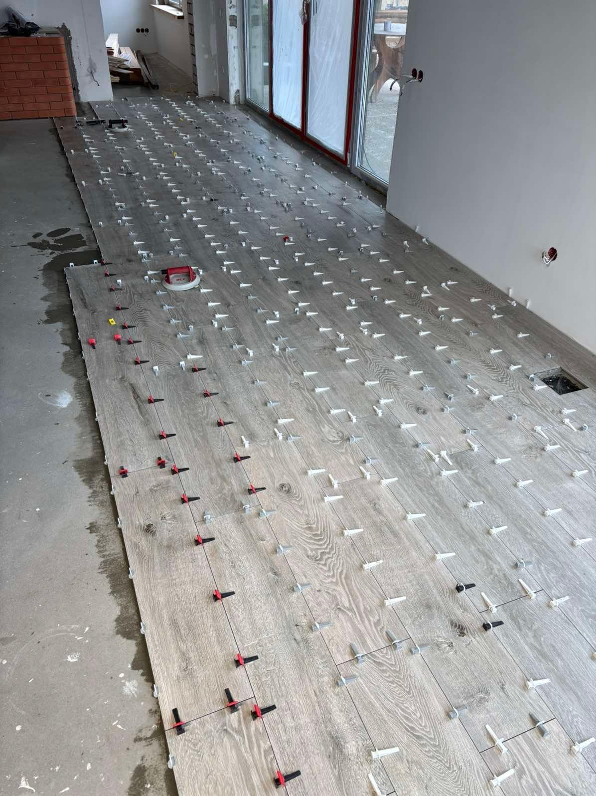 Układanie paneli podłogowych z użyciem systemu poziomowania. Widoczne białe klipsy i czerwone kliny. Częściowo ułożona podłoga w jasnym odcieniu drewna.