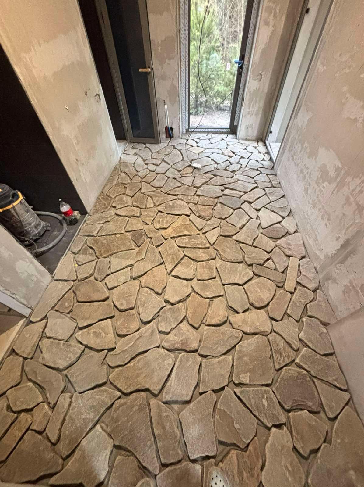 Kamienna posadzka z nieregularnych płyt w korytarzu, surowe ściany w tle. Widoczny odkurzacz i butelka. Naturalne światło z okna.