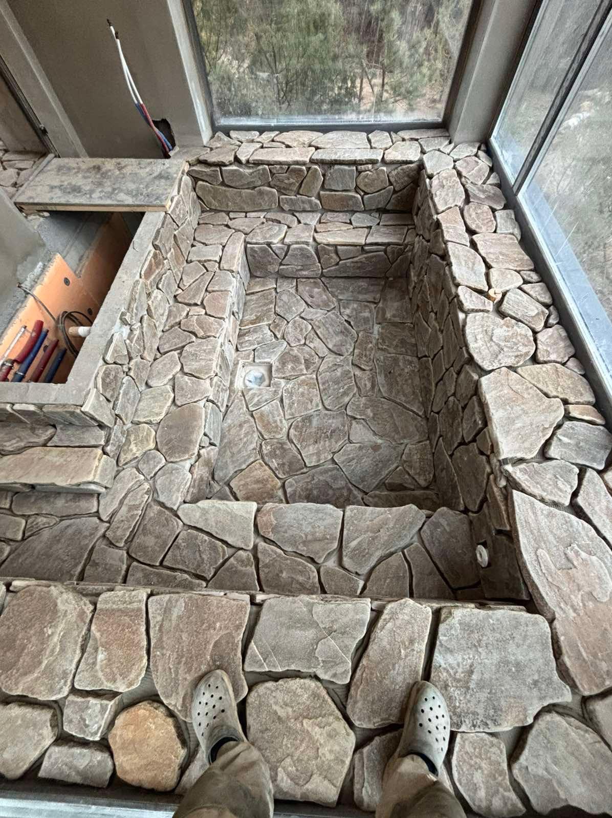 Kamienna obudowa wnęki okiennej w trakcie prac. Widoczne fragmenty instalacji i okno z widokiem na zewnątrz. U dołu stopy w obuwiu roboczym.