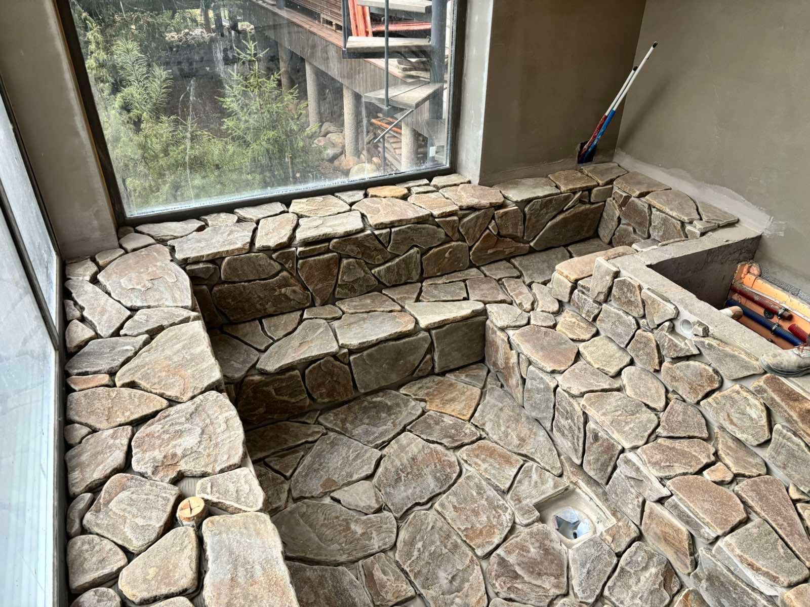 Wykończona kamieniem łazienka z dużym oknem i widokiem na zieleń. Kamienna mozaika na ścianach i podłodze, odpływ liniowy. W tle widoczne narzędzia i elementy instalacji.