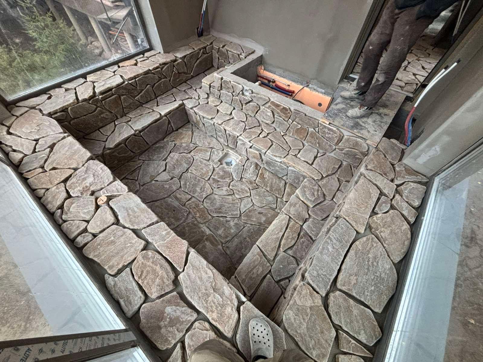 Kamienna mozaika w łazience, ułożona z nieregularnych, szarych kamieni. Widoczne okno i fragment instalacji. Narożne ujęcie podkreśla unikalny wzór i fakturę.