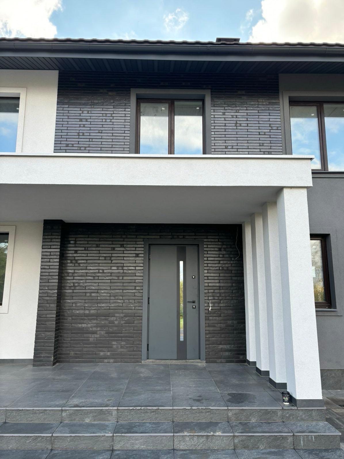 Elewacja domu z ciemnej cegły klinkierowej i szarą stolarką okienną w Redzie. Nowoczesne wykończenie z widocznymi schodami wejściowymi i kolumnami.