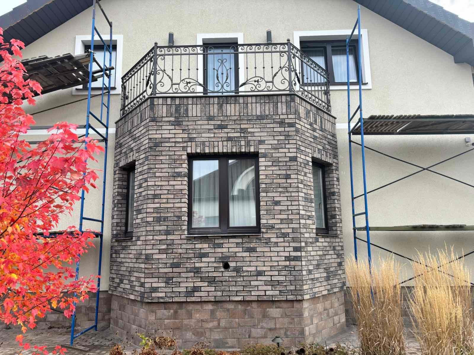 Elewacja domu z balkonem wykończona cegłą klinkierową w odcieniach szarości i beżu. Widoczne rusztowania i dekoracyjna, metalowa balustrada balkonu.