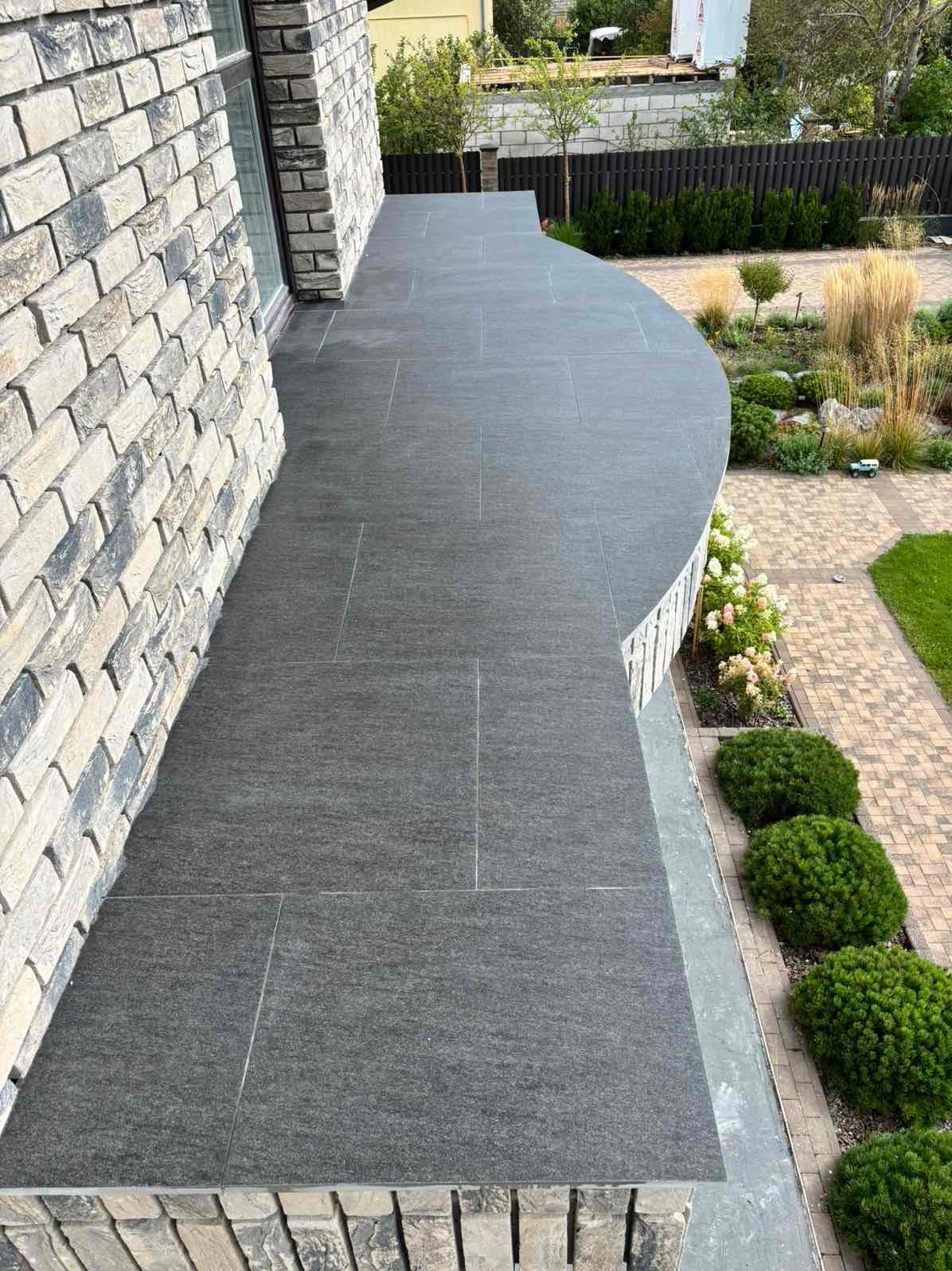 Nowoczesny balkon z szarymi płytkami, wykończony cegłą klinkierową. Widok na zadbany ogród z krzewami i ścieżką. Płytki ułożone z precyzją, tworzą gładką powierzchnię.