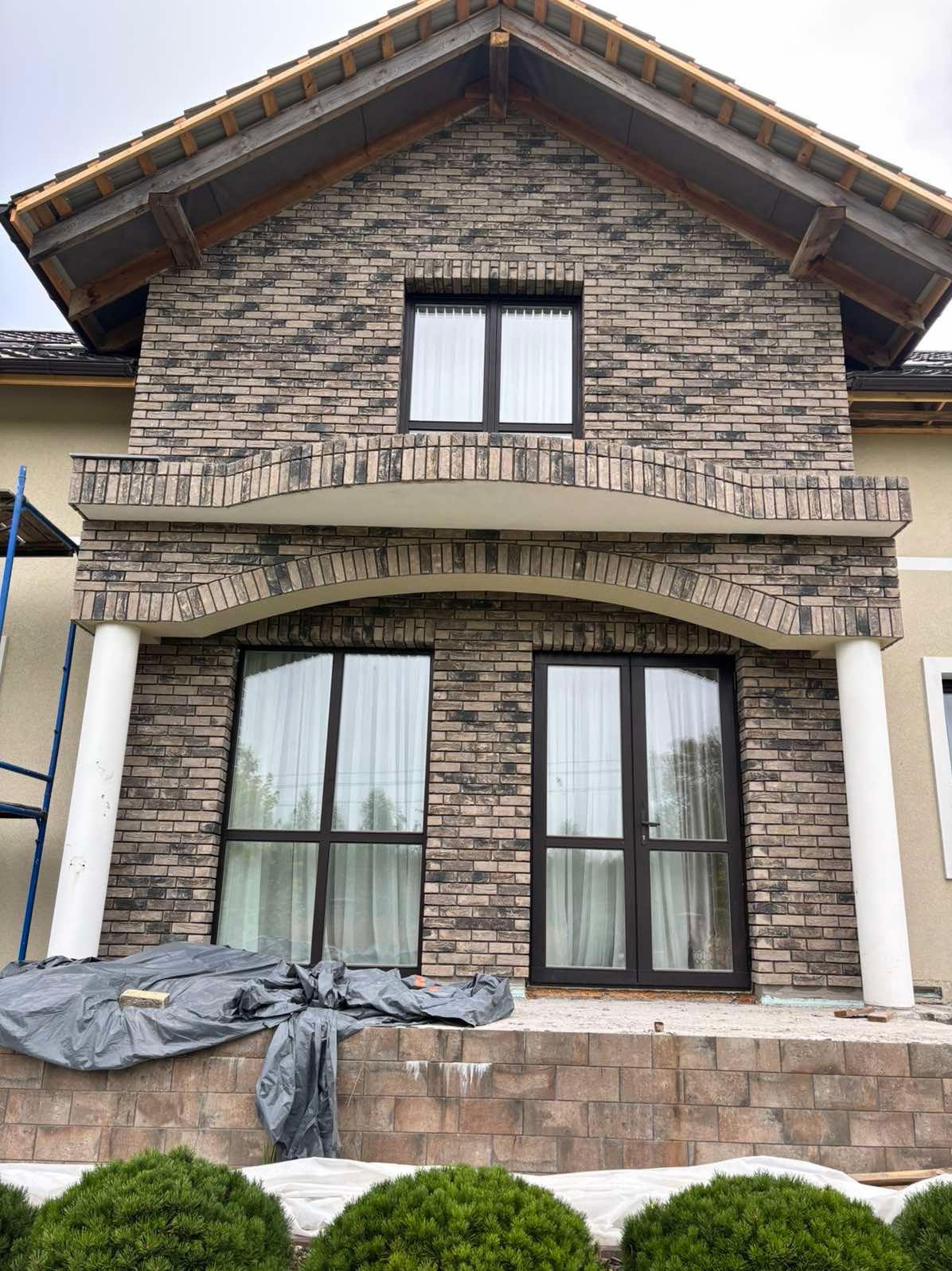 Elewacja domu z cegły klinkierowej w trakcie prac wykończeniowych. Widoczne okna z czarnymi ramami, kolumny i rusztowanie. Częściowo zakryte podłoże.