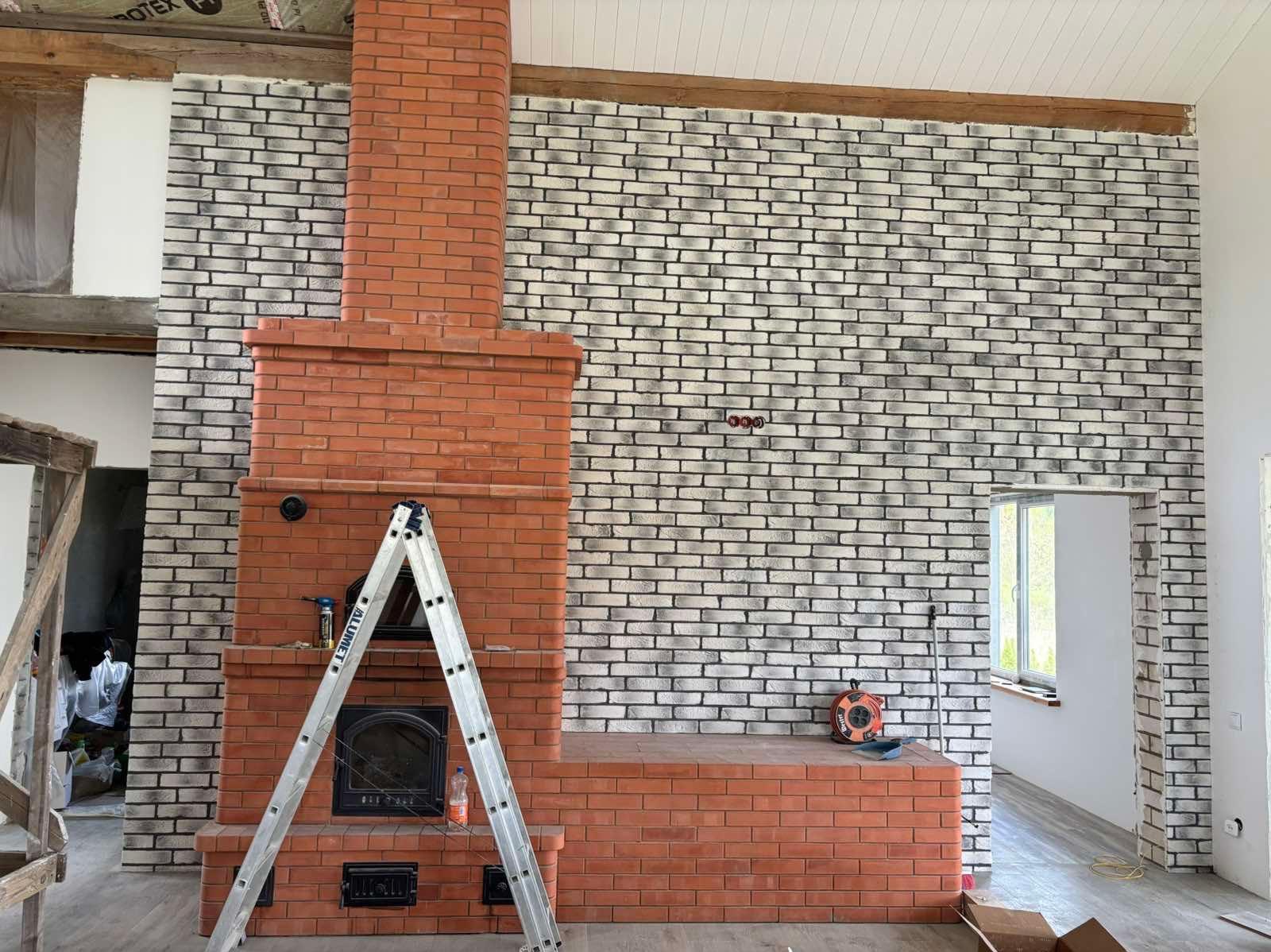 Murowany kominek z cegły i ściana z cegły klinkierowej w odcieniach szarości, drabina aluminiowa oparta o kominek, widoczne gniazdka elektryczne na ścianie.