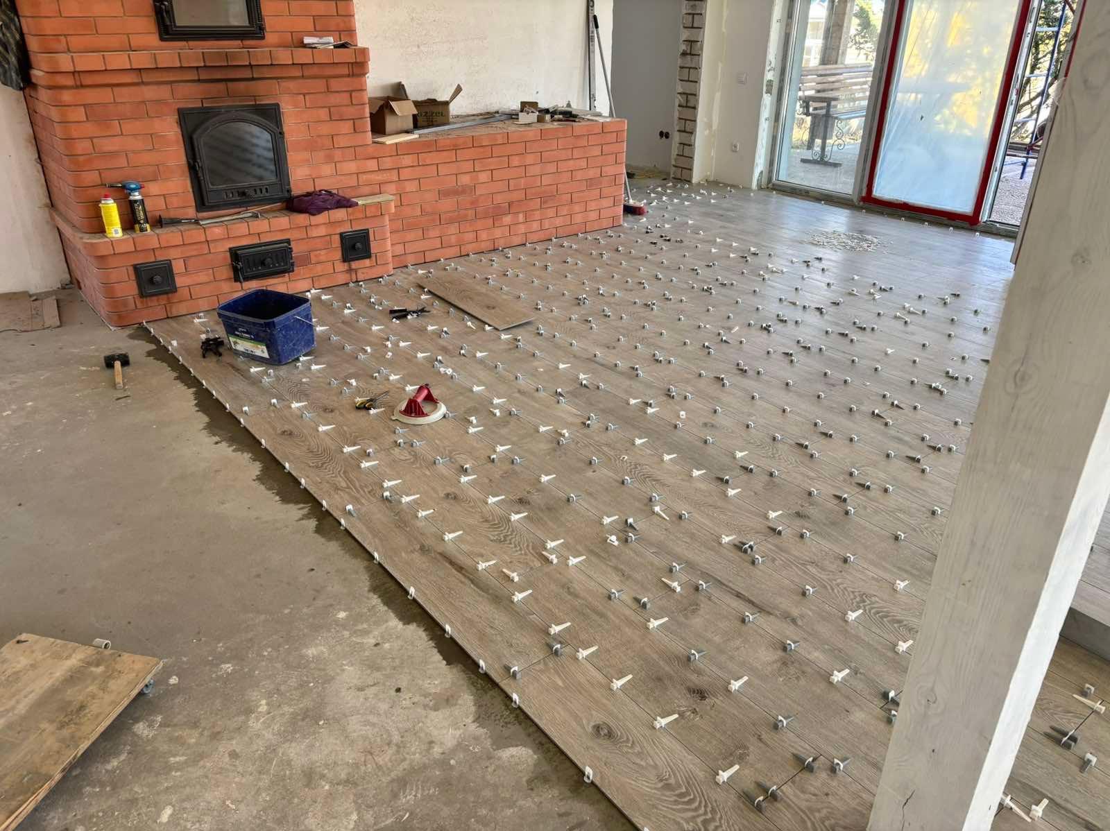 Układanie paneli podłogowych w dużym pokoju z ceglanym kominkiem. Widoczne narzędzia i system poziomowania paneli. Jasne panele z wyraźną strukturą drewna.