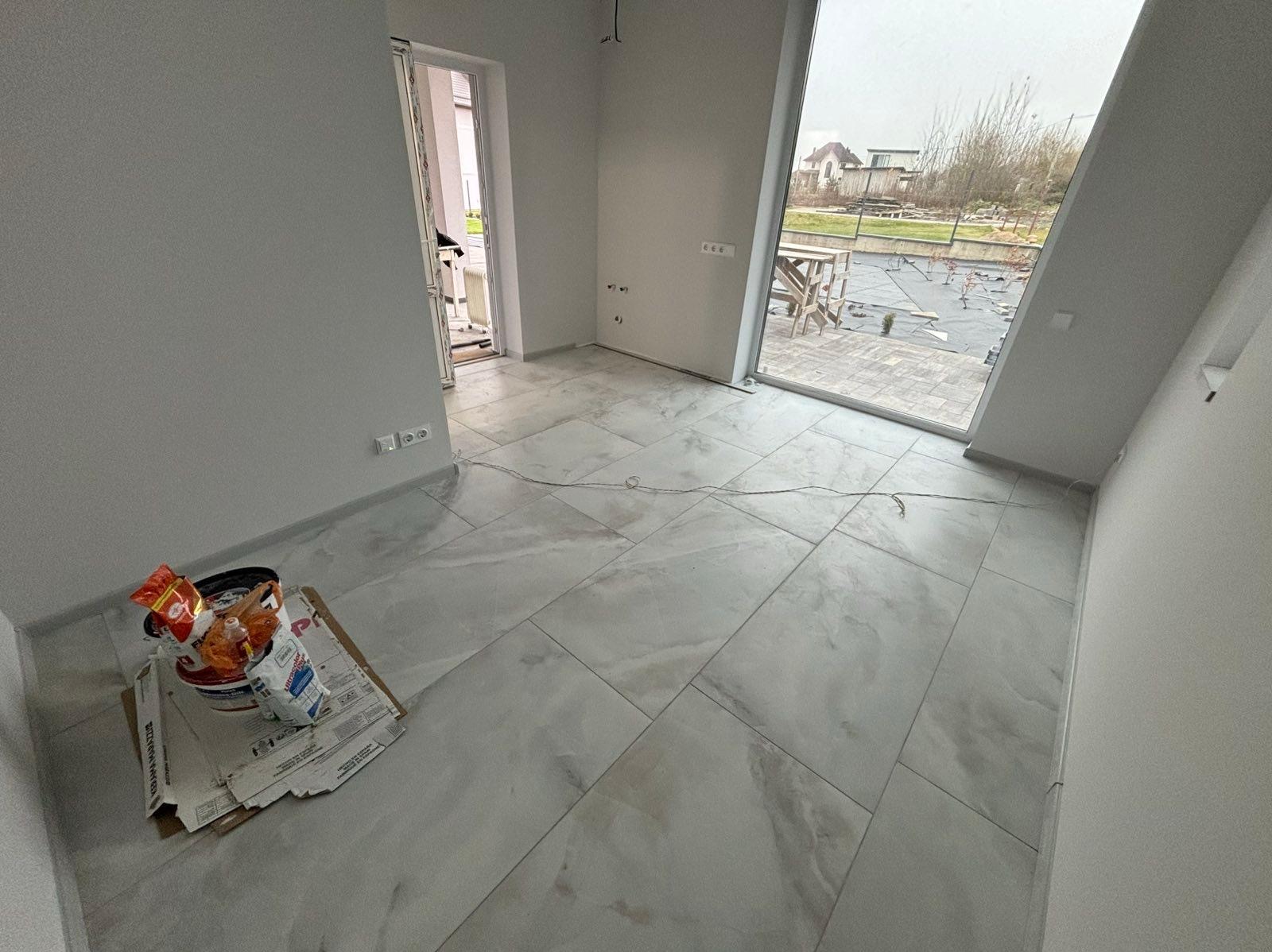 Jasne, marmurowe płytki podłogowe w nowym wnętrzu. Widoczne wiaderka z zaprawą i narzędzia. Duże okno z widokiem na zewnątrz. Minimalistyczny styl wykończenia.