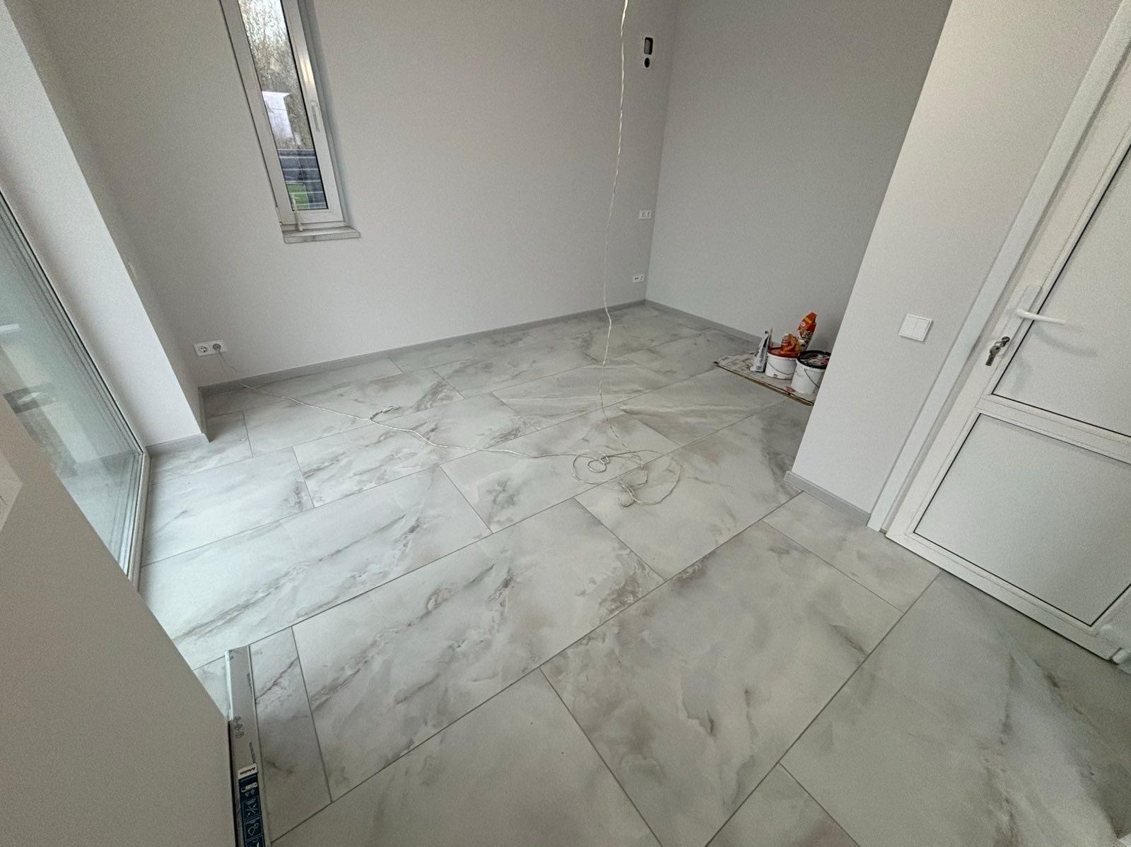 Jasne, marmurowe płytki podłogowe w nowym pokoju z oknem i białymi ścianami. Widoczne wiaderka z farbą i kabel zwisający z sufitu. Minimalistyczny design.