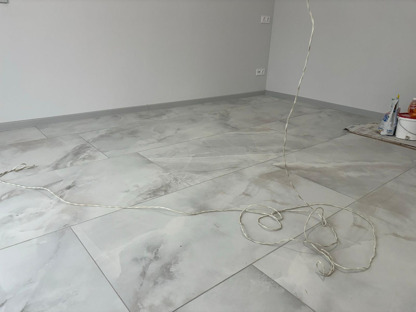 Jasne, marmurowe płytki podłogowe w nowym pomieszczeniu z szarą listwą przypodłogową i zwisającym przewodem. Widoczne gniazdka elektryczne i akcesoria malarskie.