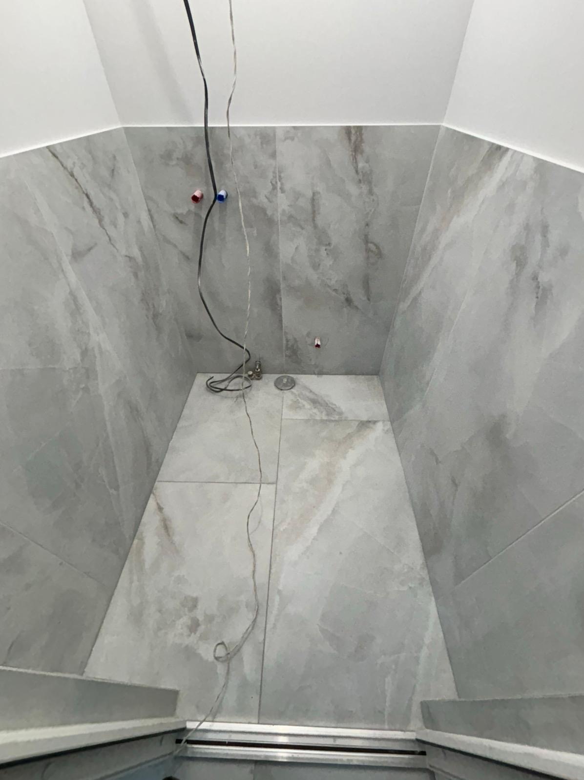 Wnętrze kabiny prysznicowej w trakcie prac. Widoczne szare płytki imitujące marmur na ścianach i podłodze, wystające kable i rury. Etap przed montażem armatury.