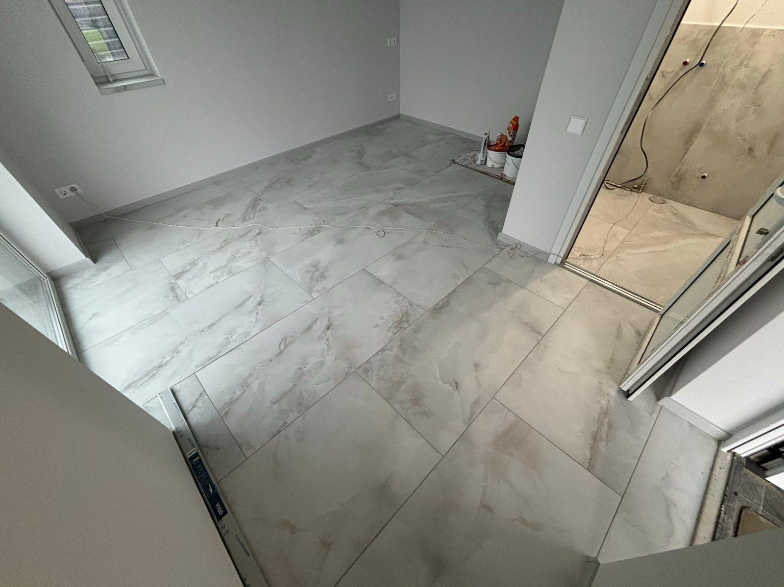 Wykończona łazienka z płytkami imitującymi marmur na podłodze i ścianach kabiny prysznicowej. Widoczne akcesoria glazurnicze. Minimalistyczny styl.