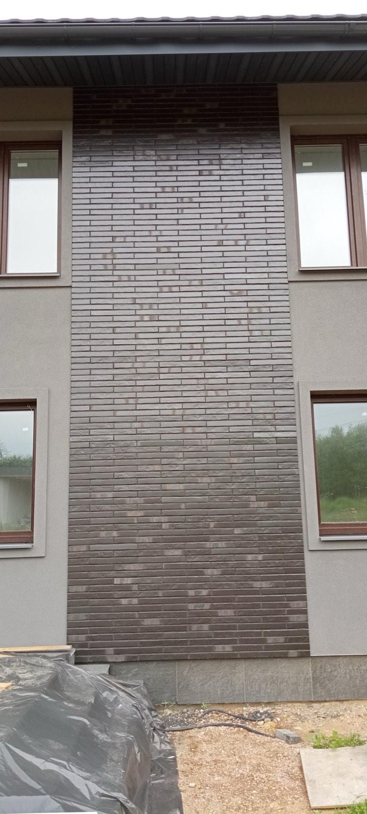 Elewacja budynku z brązową cegłą klinkierową, widoczne okna z brązowymi ramami. Część terenu zakryta folią budowlaną. Detal wykończenia zewnętrznego.