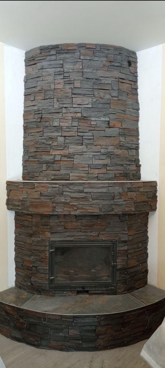 Obudowa kominka z kamienia dekoracyjnego w odcieniach szarości i brązu, ułożona w poziome pasy. Widoczny fragment ściany i podłogi. Całość w neutralnym oświetleniu.