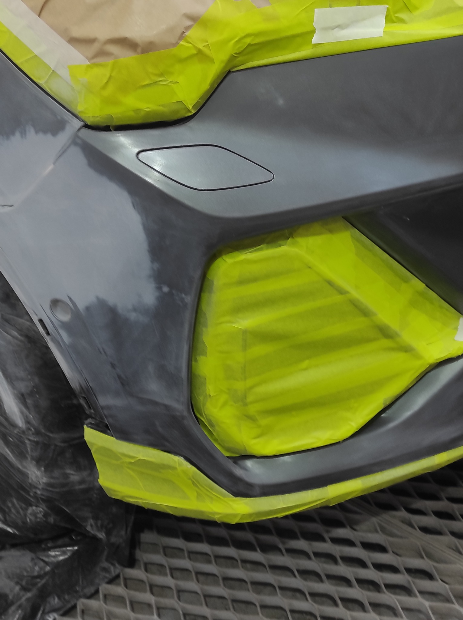 Przygotowany do lakierowania fragment karoserii auta, zabezpieczony żółtą taśmą maskującą. Widoczne ślady szpachlowania i podkładu. Element na metalowej kratownicy.