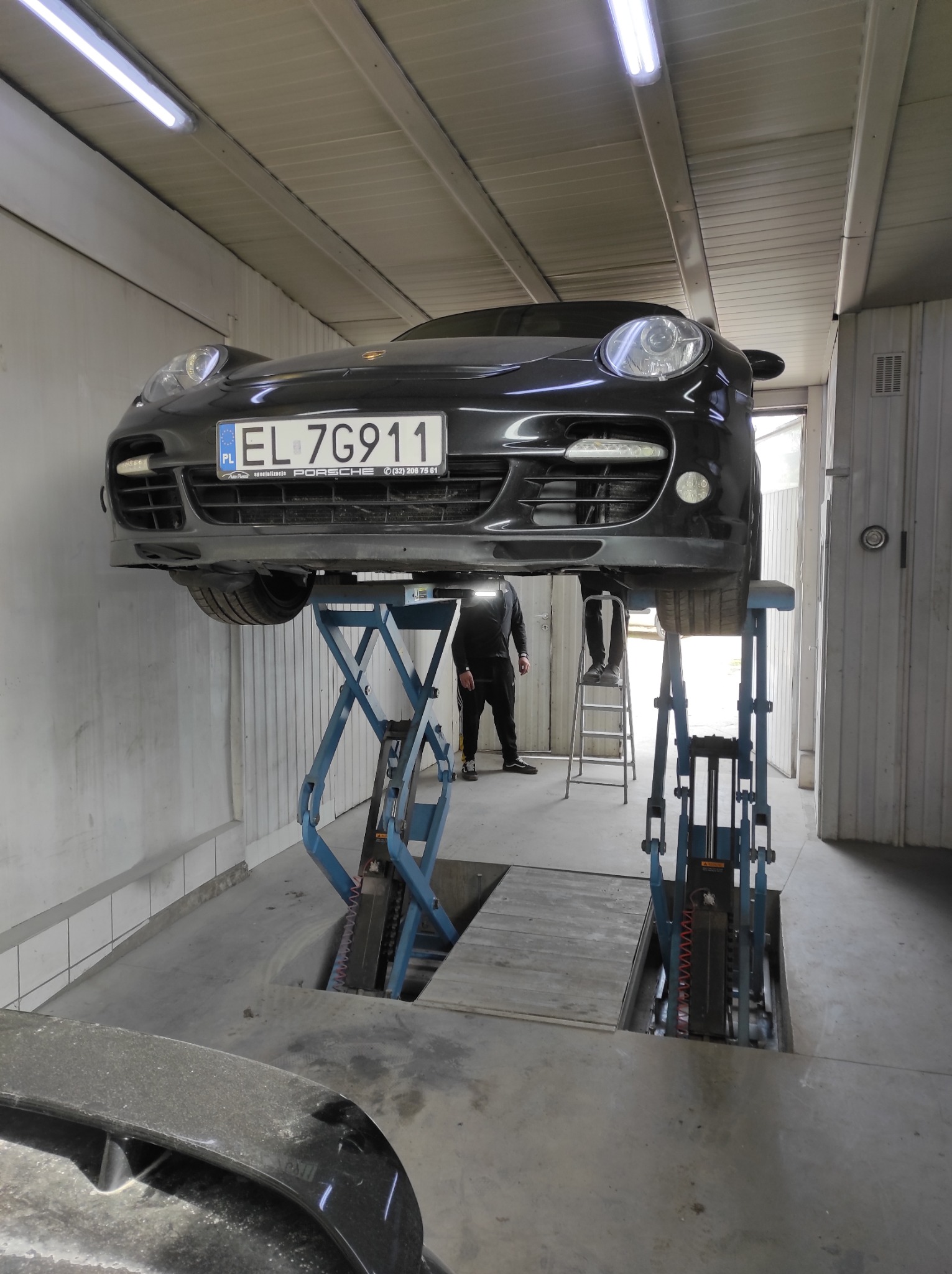 Czarny Porsche na podnośniku w warsztacie. Widoczny numer rejestracyjny EL 7G911 i pracownik na drabinie. Oświetlenie jarzeniowe.