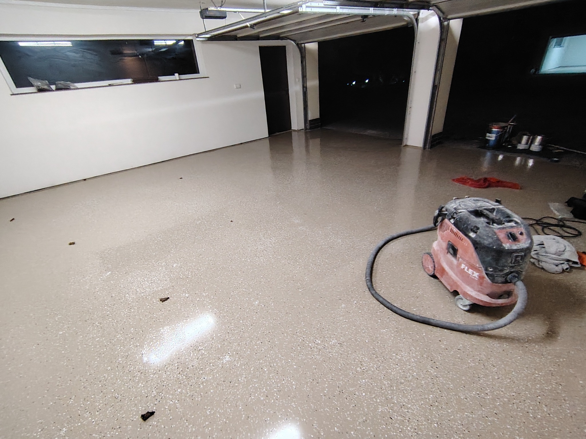 Błyszcząca, beżowa posadzka z drobnymi plamkami w garażu, z widocznym odkurzaczem przemysłowym Flex. W tle otwarte wrota garażowe i okno.