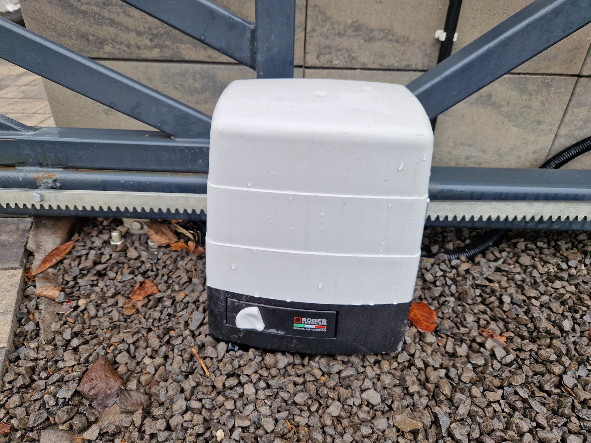 Zamknięty napęd bramy przesuwnej Roger Digital Automation, zamontowany na szarej bramie, na tle szarej kostki brukowej i kamieni. Widoczne krople wody.