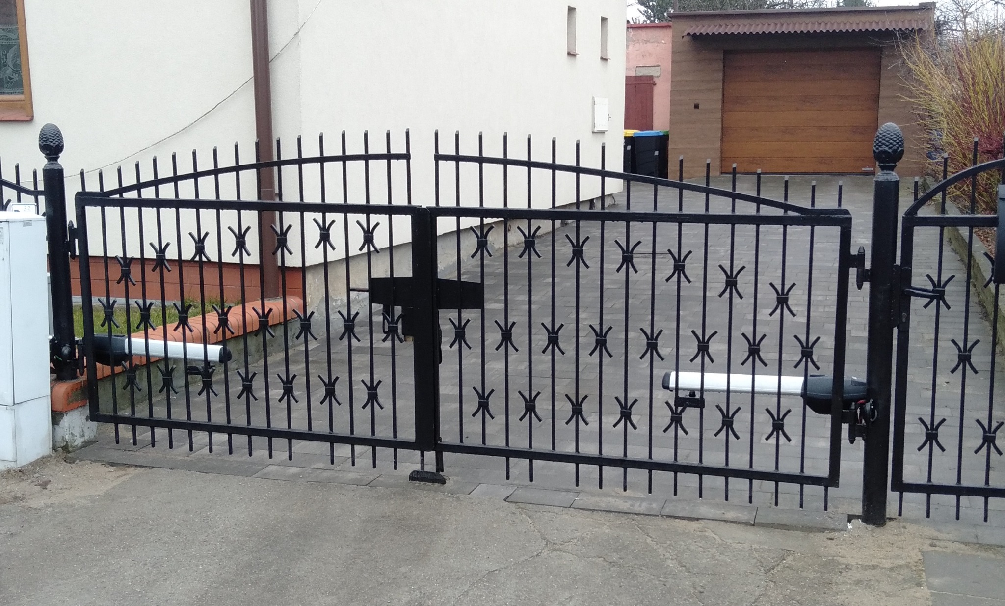 Czarna, metalowa brama wjazdowa z automatyką, widoczna na tle domu i garażu. Dekoracyjne elementy w kształcie gwiazdek. Napęd bramy z białymi siłownikami.