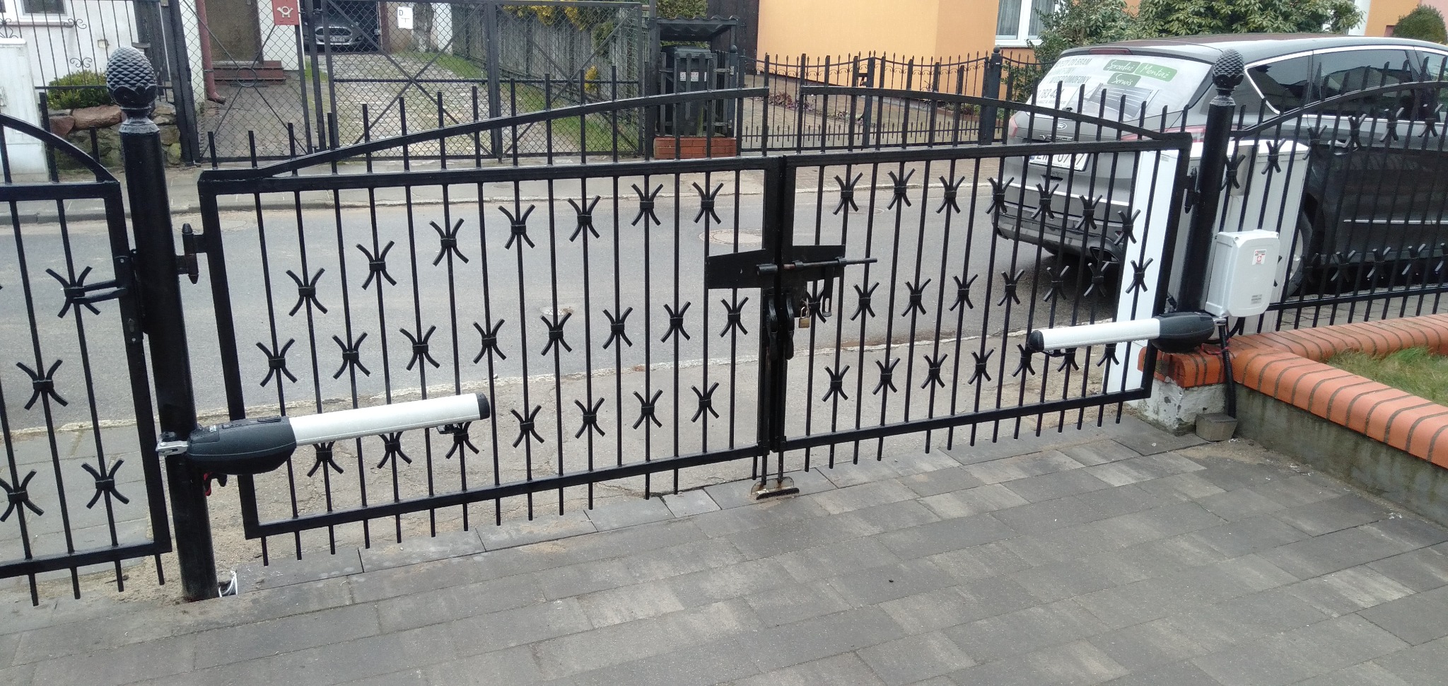 Czarna, metalowa brama wjazdowa z automatyką, widoczna z zewnątrz posesji. Brama ozdobiona motywem krzyżyków, z zamkiem na środku i siłownikami po bokach.