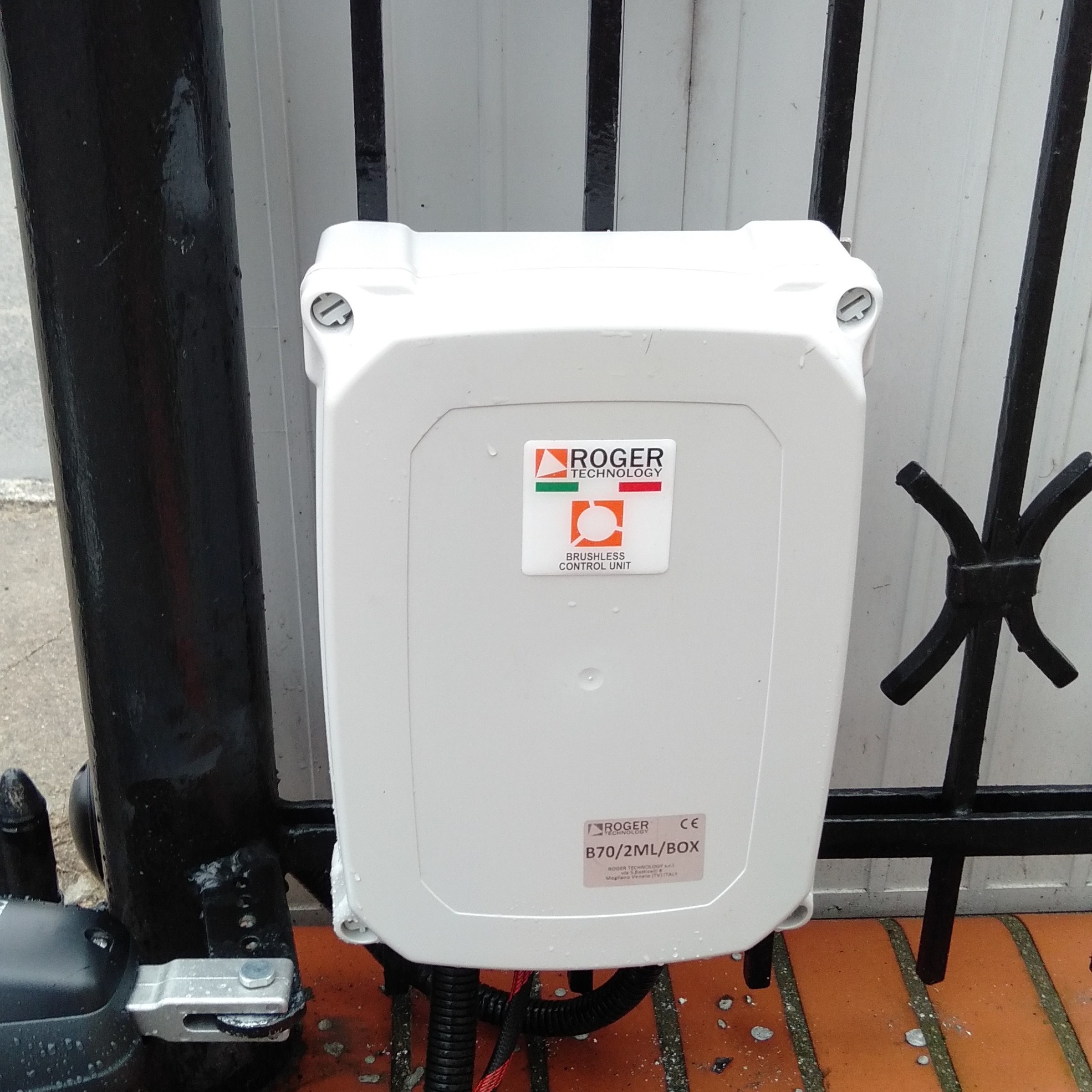 Biała skrzynka sterownicza Roger Technology zamontowana na czarnej, metalowej bramie wjazdowej. Widoczne przewody i fragment napędu. Ceglana podstawa.