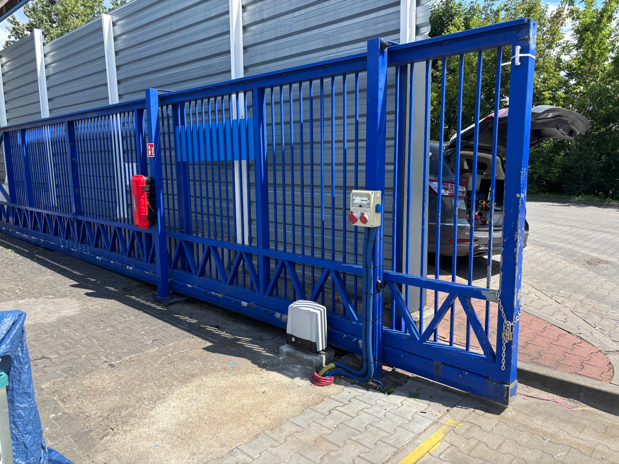 Niebieska brama przesuwna z automatyką i skrzynką elektryczną na zewnątrz, na tle szarego ogrodzenia i samochodu z otwartym bagażnikiem.