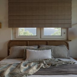 Totemi Marta Kowalska - Sypialnia w neutralnych barwach: wezgłowie z plecionki, drewniana szafka nocna, tekstylna roleta rzymska i lampki ścienne z abażurami. Okno z widokiem na zieleń.