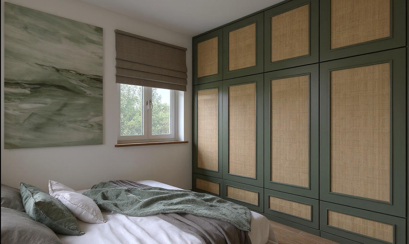 Sypialnia w beżach i zieleni: łóżko z dekoracyjną narzutą, obraz nad wezgłowiem, okno z roletą rzymską i duża szafa z rattanowymi frontami w ciemnozielonej ramie.