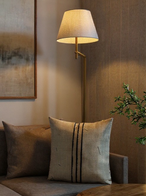 Beżowa sofa z dekoracyjnymi poduszkami w salonie, oświetlona lampą podłogową. Na ścianie obraz w drewnianej ramie. W tle widoczna ściana z teksturą.