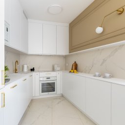Howesto Sp z o.o. - Biała kuchnia z marmurowym blatem i złotymi akcentami: bateria, uchwyty, lampa. Minimalistyczny design, jasne wnętrze, dwie filiżanki na blacie.