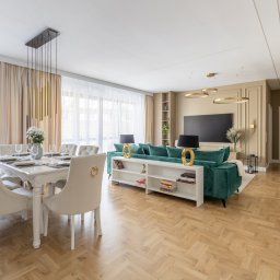 Howesto Sp z o.o. - Eleganckie wnętrze: parkiet w jodełkę, stół z nakryciem, krzesła z ozdobnymi uchwytami, zielona sofa i złote lampy. Całość w jasnych, stonowanych barwach.