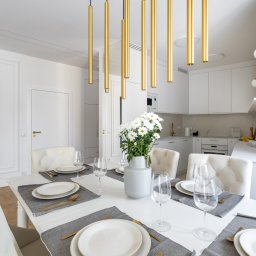 Howesto Sp z o.o. - Jasne, nowoczesne wnętrze jadalni z białym stołem, krzesłami i złotymi akcentami oświetleniowymi. W tle widoczna kuchnia z białymi szafkami i marmurowym blatem.
